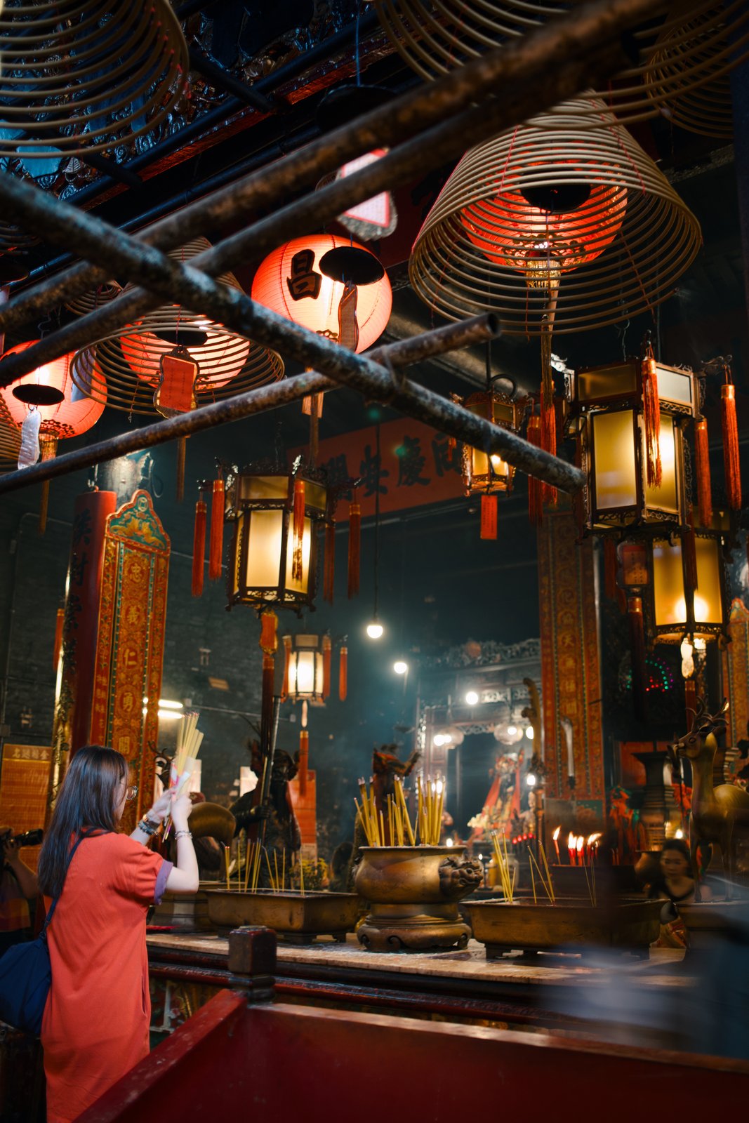 A woman lifting incense sticks beneath hanging coils and lanterns in a temple